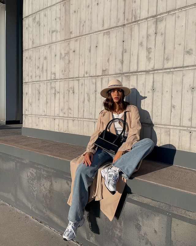 Foto de uma mulher sentada em uma calçada alta. Ela usa uma camiseta branca, sobretudo longo bege, calça jeans, tênis branco esportivo, bolsa preta e chapéu bege. Ela olha para a câmera e não sorri.