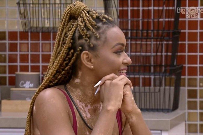 Take da participante do BBB22, Natália. Ela é negra, está com cabelo com box braids e está sentada com os braços apoiados na mesa.