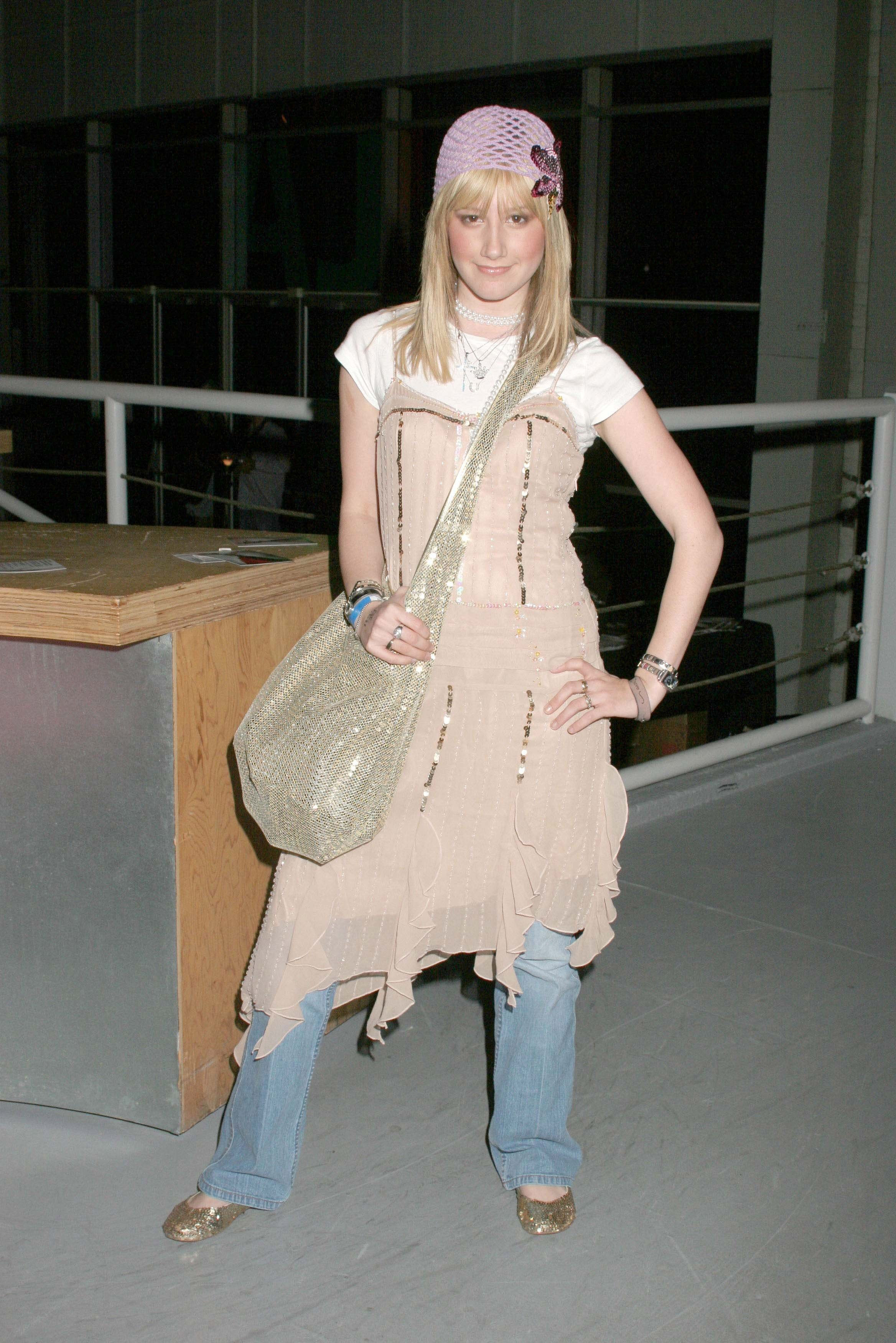 ashley-tisdale-look-vestido-com-calca Foto da atriz Ashley Tisdale. Ela usa um vestido com babados rosa claro, calça jeans, sandália e bolsa de ombro bege. Ela olha para a câmera e sorri levemente.