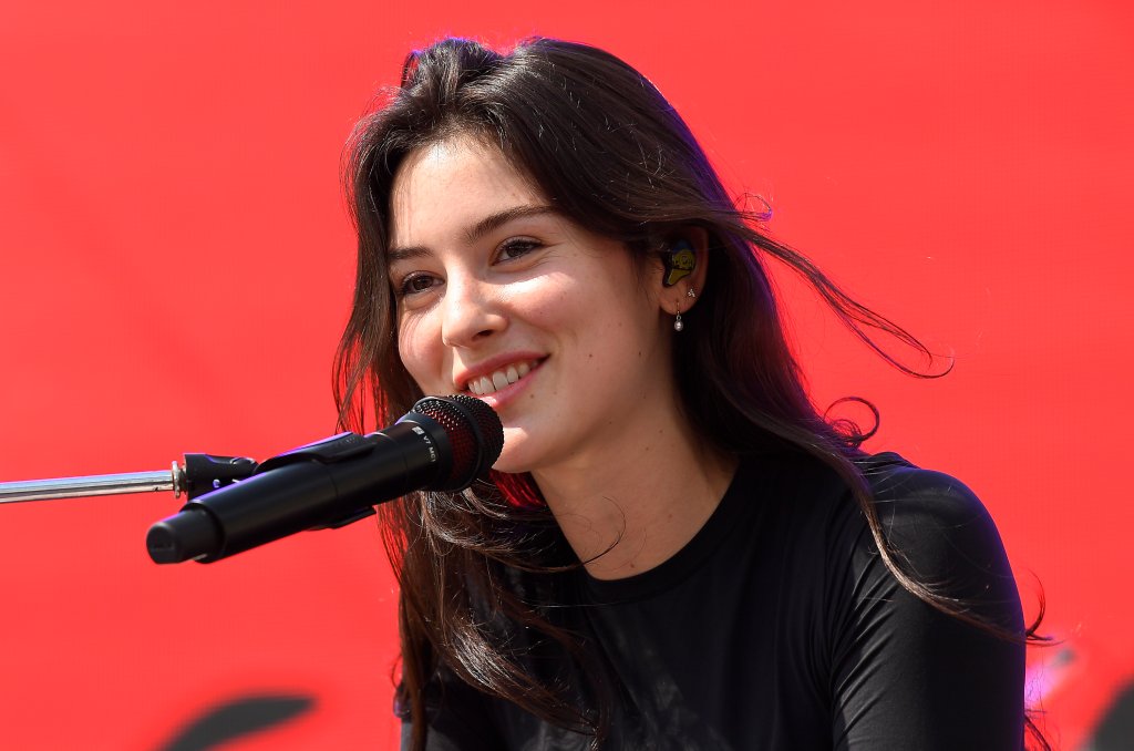 Gracie Abrams usando blusa preta e sorrindo enquanto canta em um microfone também preto com um telão vermelho ao fundo