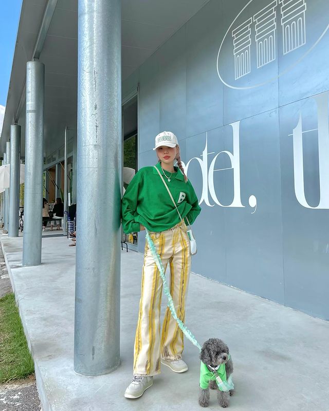look-com-moletom-verde-bottega Foto de uma mulher em uma calçada. Ela usa um boné branco, bolsa branca, moletom Verde Bottega, calça amarela e tênis branco. Ela segura a coleira de um cachorro com a mão direita, olha para a câmera e sorri levemente.