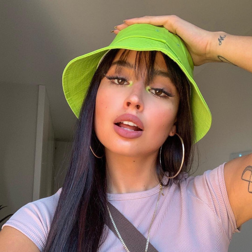 Selfie de uma mulher. Ela usa um bucket hat verde, cabelo solt, brinco de argola, camiseta lilás e maquiagem com delineador e ponto de luz neon no canto interno dos olhos. Ela está com a mão esquerda na cabeça, olha para a câmera e não sorri.