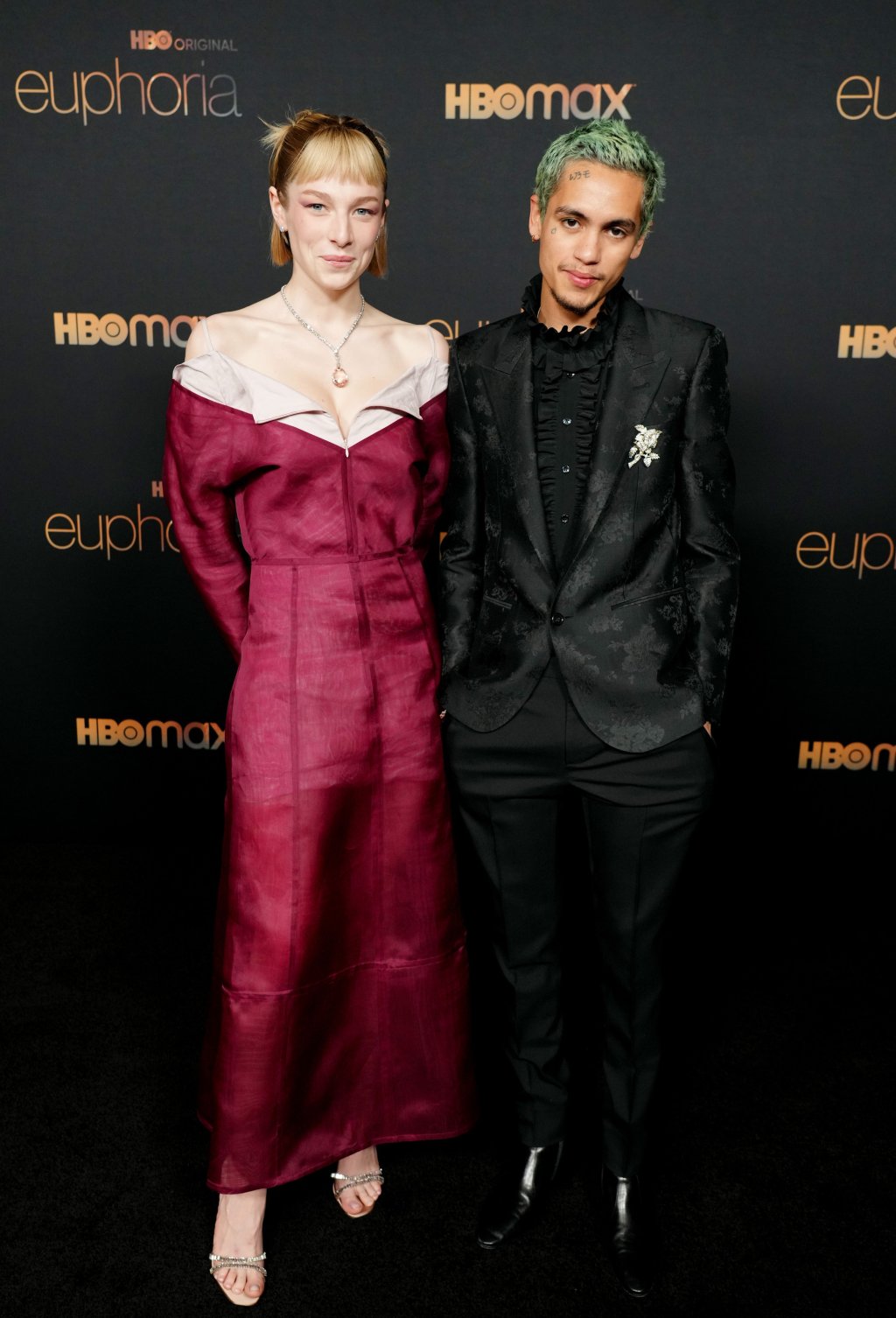 Hunter Schafer e Dominic Fike sorrindo levemente enquanto posam para foto na premiere da 2ª temporada de Euphoria; ela veste uma roupa rosa com detalhe branco e ele usa terno e camisa na cor preta; os dois estão com as mãos no bolso