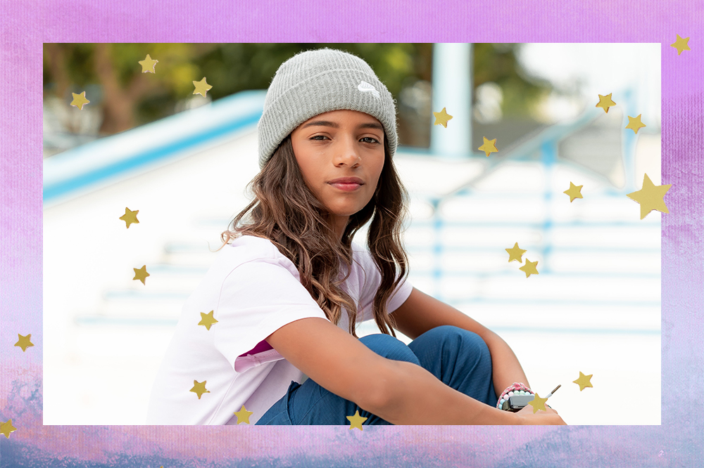 Rayssa Leal usando gorro, camiseta branca e calça azul em fundo lilás, roxo e azul com estrelinhas douradas