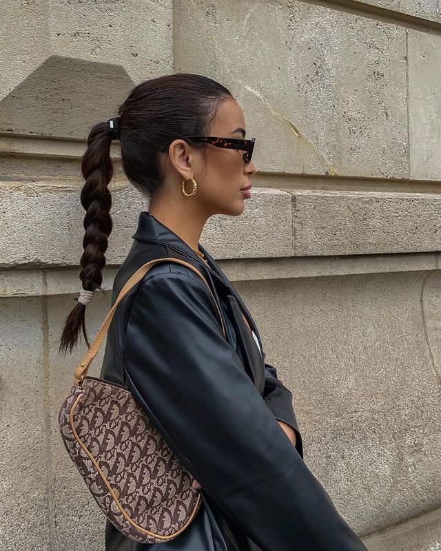 trança no rabo de cavalo Foto de uma mulher de perfil em uma calçada. Ela usa uma jaqueta de couro preta, bolsa baguete marrom, óculos de sol preto, brinco de argola dourado e penteado rabo de cavalo alto trançado. Ela olha para frente e não sorri para a foto.