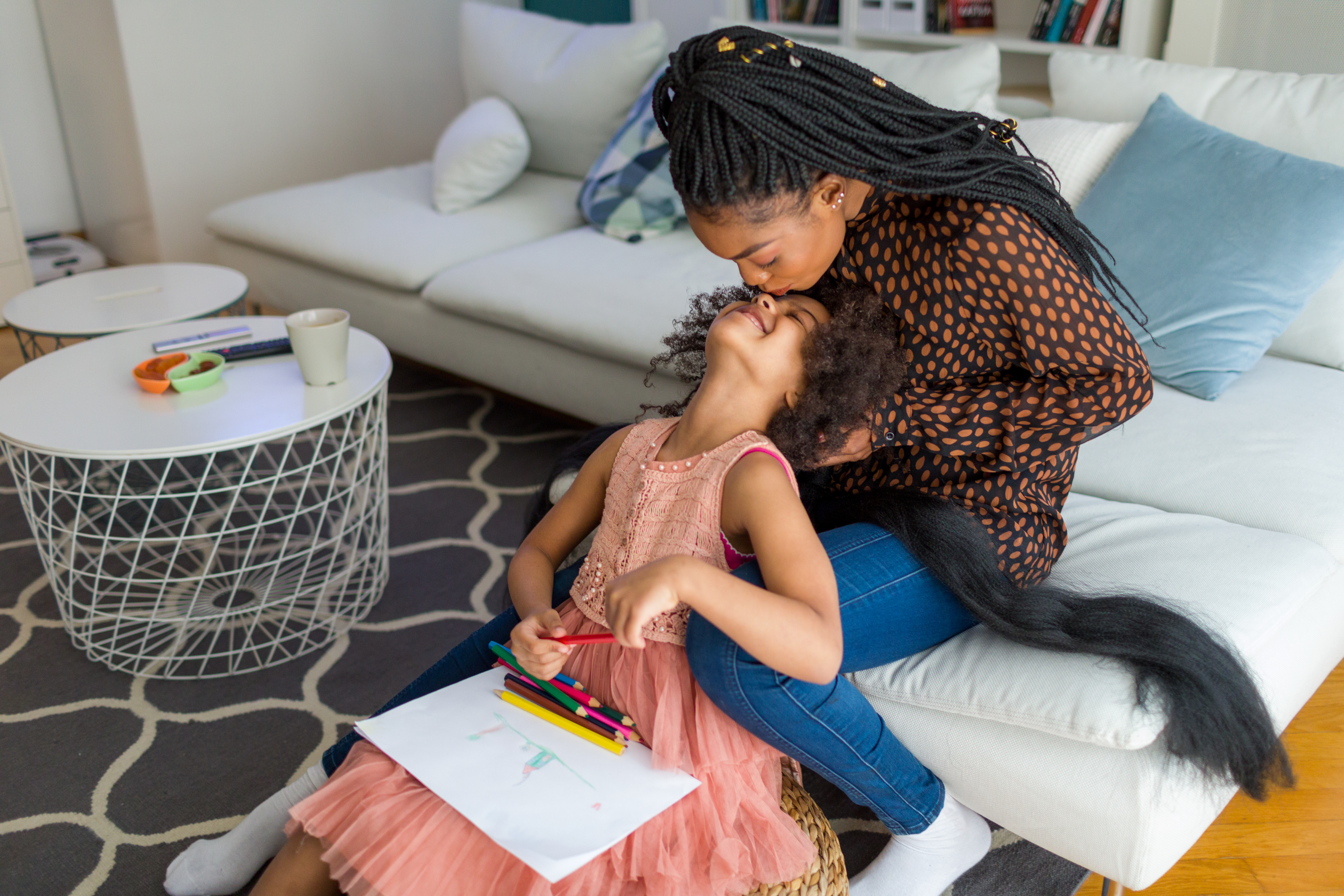 African Mother Braiding Her Daughter’s Hair At Home Imagem mostra mãe e filha sentadas no sofá e no chão, respectivamente. Ambas então em uma sala de estar enquanto a mãe trança o cabelo da filha e a mesma olha para cima e recebe um beijo na testa. O registro de trocas e afetos entre mãe e filha segue acompanhado de uma borda colorida e estrelinhas douradas por toda a imagem.