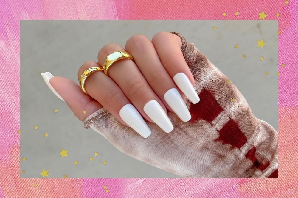 Montagem com o fundo rosa e detalhe de estrelinhas douradas com a foto de uma mão com as unhas longas esmaltadas de branco e anéis dourados no dedo indicador e médio.