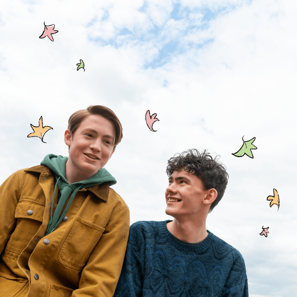 Nick e Charlie, da adaptação de Heartstopper; Nick está olhando pra frente e sorrindo enquanto Charlie olha para ele também sorrindo; ao fundo é possível ver o céu azul com nuvens brancas e folhas ilustradas nas cores rosa, amarelo e verde