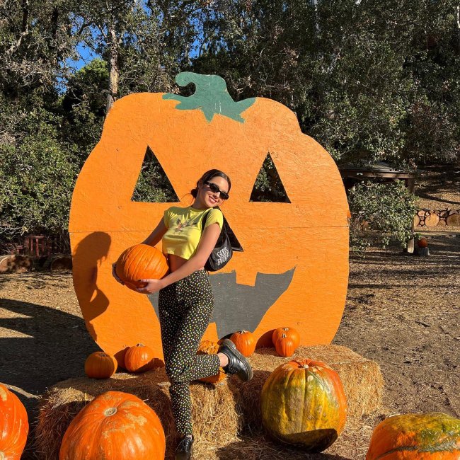 Foto da cantora Olivia Rodrigo em um local aberto com abóboras no chão. Ela usa uma baby tee amarela, calça estampada e calça um oxford. Ela segura uma abóbora, olha para a câmera e sorri.