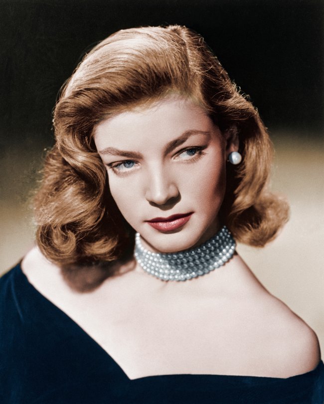 Retrato da atriz Lauren Bacall nos anos 40. Ela usa um vestido ombro a ombro preto, colar de pérolas, brinco de pérola, cabelo solto e maquiagem simples. Ela olha para o lado e não sorri.