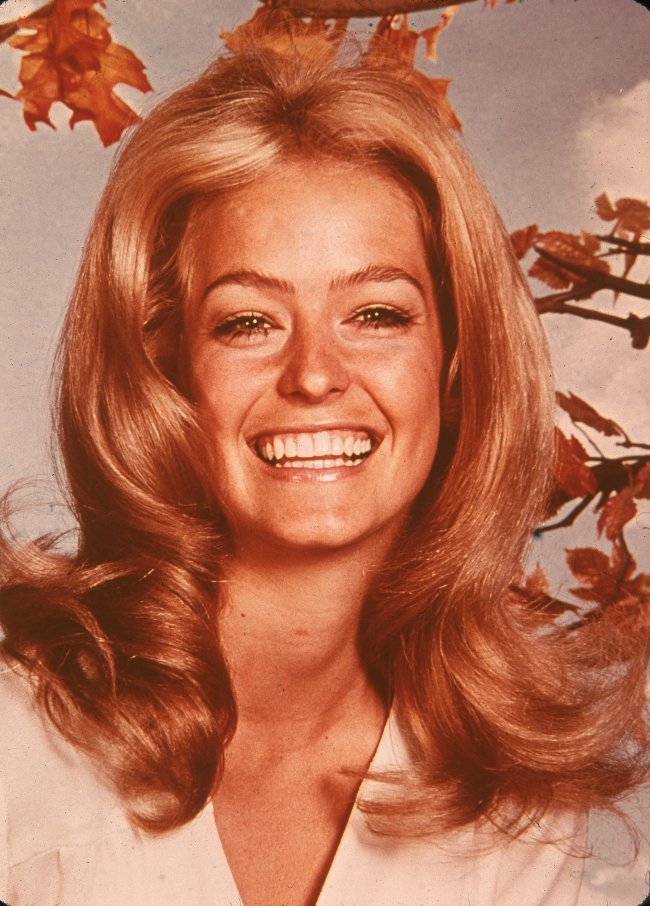 Foto da atriz Farrah Fawcett nos anos 70. Ela está com uma camiseta branca, cabelo solto e maquiagem leve. Ela olha para a câmera e sorri.