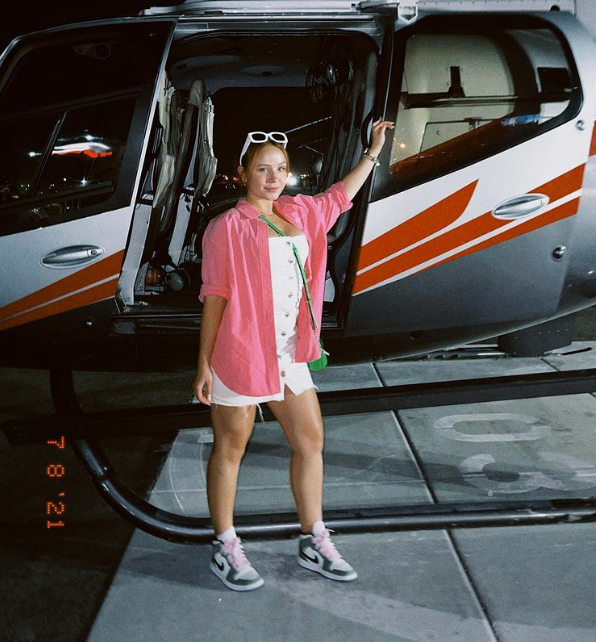 Larissa Manoela usando look com vestido branco, camisa oversized rosa, tênis, bolsa transpassada verde e óculos de sol brancos na cabeça. Ela está em frente a um helicóptero