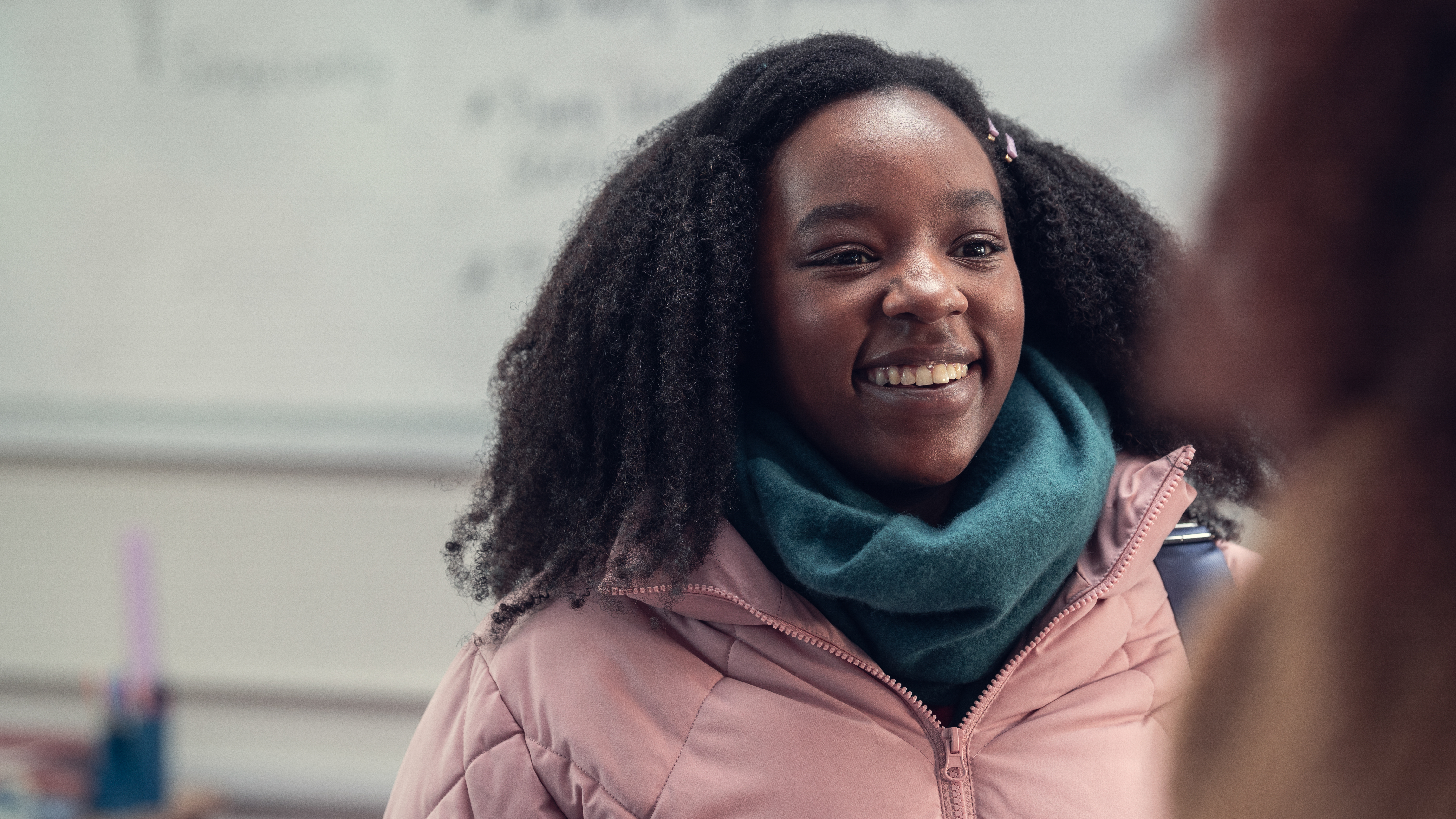 Tara Jones (Corinna Brown) em Heartstopper. Ela está sorrindo, usando casaco rosa e cachecol azul