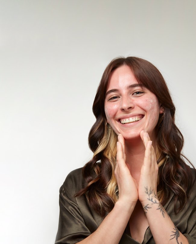 encontre-o-melhor-tratamento-para-a-sua-pele Foto de uma modelo com em um lugar com fundo branco, blusa preta, cabelo solto e pele limpa. Ela está com as duas mãos no rosto aplicando um produto, olha para a câmera e sorri.