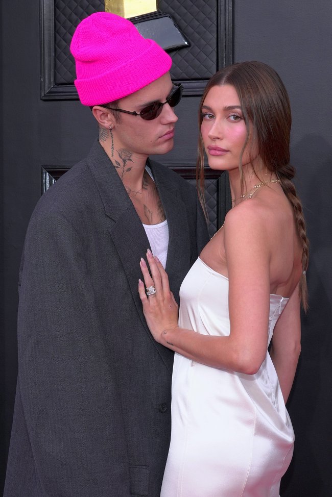 Foto Justin e Hailey Bieber no tapete vermelho do Grammy 2022. Na imagem Justin usa uma touca rosa, blazer oversized cinza, regata branca e óculos escuros. Hailey usa um vestido branco tomara que caia, maquiagem natural e cabelo preso em uma trança boho despojada.