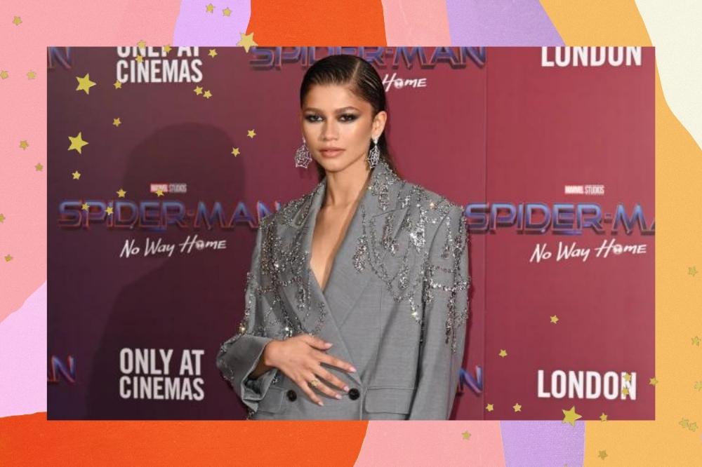 Montagem com o fundo colorido e detalhe de estrelas douradas nas bordas com a foto da Zendaya no centro. Na foto ela está na photocall de Homem-Aranha: Sem Volta para Casa, em Londres usando blazer oversized cinza com meia-calça preta, ambos com aplicações de cristais que imitam teia de aranha