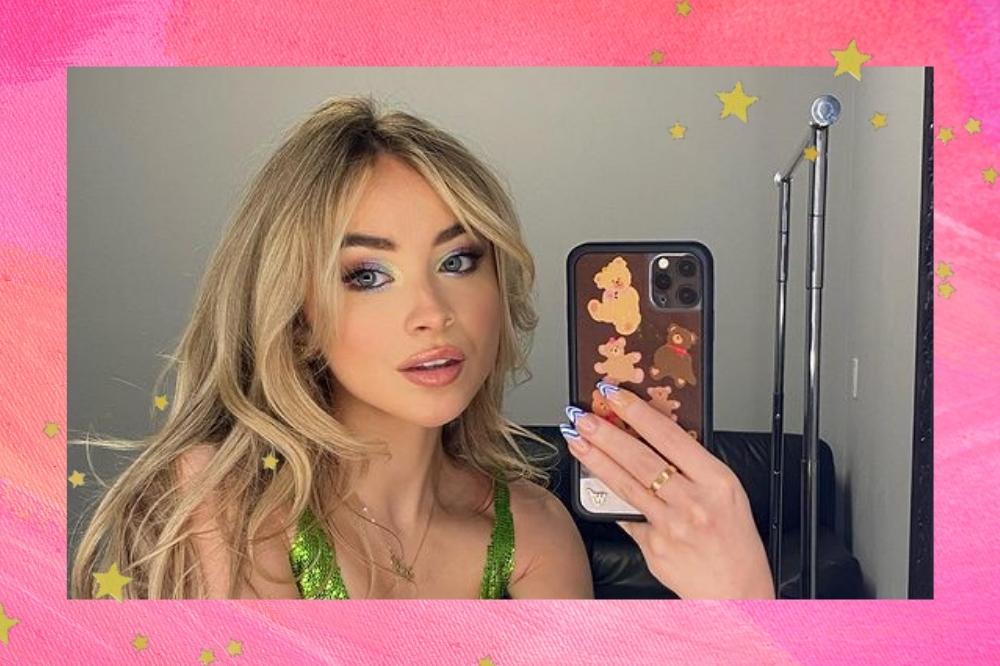 Montagem com o fundo rosa e detalhe de estrelas douradas nas bordas com a foto da atriz e cantora Sabrina Carpenter no centro. A imagem de Sabrina é uma selfie no espelho, ela usa um vestido de alcinha verde, cabelo solto e maquiagem com pele natural e olho colorido em tons pastel. Ela olha para o espelho e não sorri.