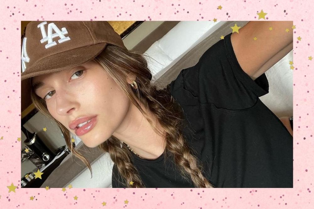 Montagem com o fundo rosa e detalhe de estrelas coloridas com a foto da Hailey Bieber no centro. A foto é uma selfie da modelo e ela usa uma camiseta preta, boné marrom, cabelo com tranças despojadas e está com o rosto sem maquiagem. Ela olha para a câmera e não sorri.