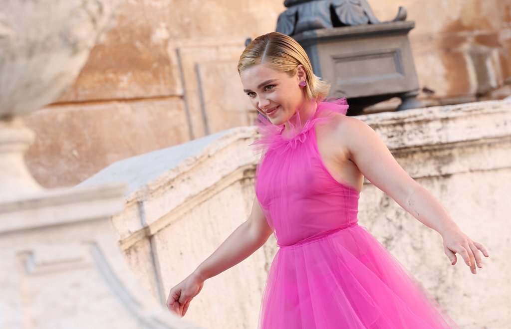 Florence Pugh sorri enquanto posa para fotos com vestido pink transparente. Ela é uma mulher branca e loira