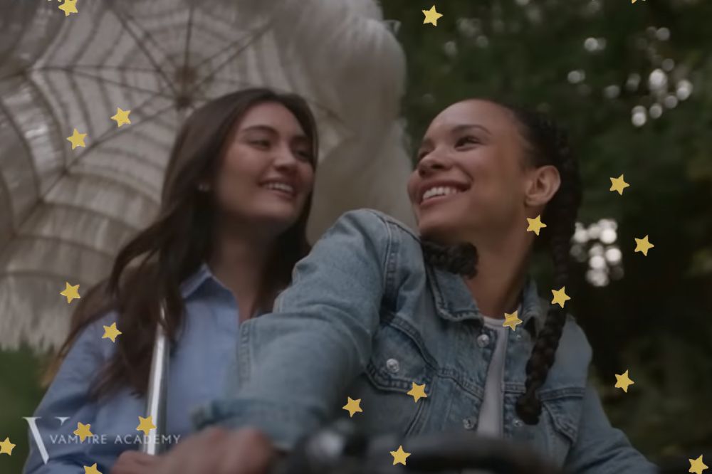 Lissa e Rose, em Vampire Academy; elas estão sorrindo e andando de bicicleta juntas; Lissa segura um guarda-chuva enquanto Rose está comandando a bicicleta