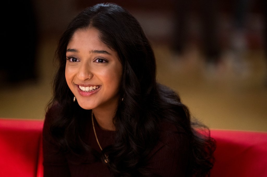 Maitreyi Ramakrishnan como Devi em Eu Nunca..., série da Netflix; ela está sorrindo e usa uma blusa vinho com um colar dourado; o fundo da imagem está desfocado