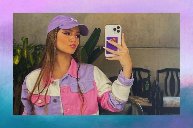 mel-maia-maquiagens-da-atriz-para-voce-se-inspirar Montagem com o fundo em degradê em tons de roxo e azul com uma selfie no espelho da atriz Mel Maia no centro. Na imagem ela usa um casaco colorido em tons de rosa, roxo e branco, boné roxo, cabelo solto e maquiagem colorida. Ela olha para o celular e faz um bico.