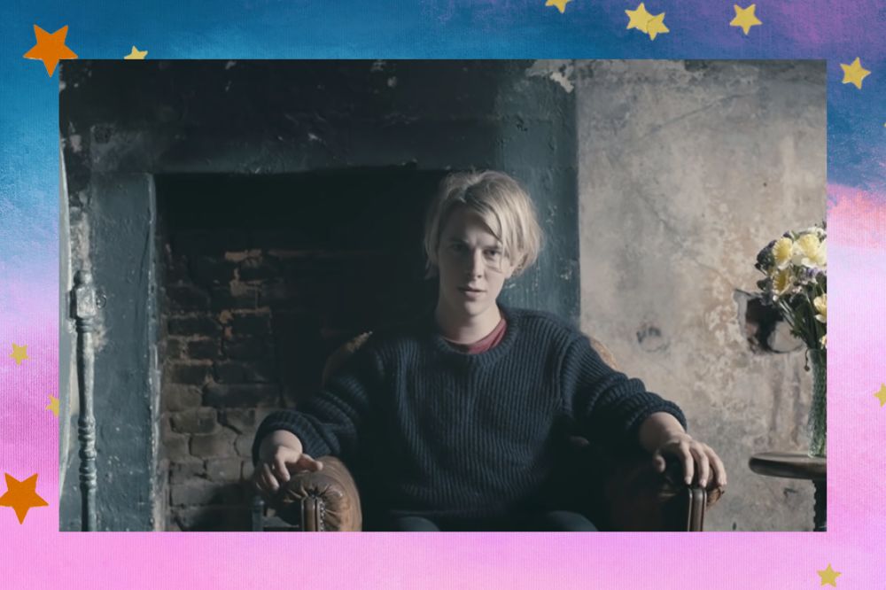 Tom Odell com expressão séria sentado em um ambiente com uma lareira ao fundo e flores em um vaso do lado direito da imagem; a margem é uma textura em degrade nas cores azul, roxo, lilás, branco e rosa com estrelas amarelas e laranjas decorando a imagem