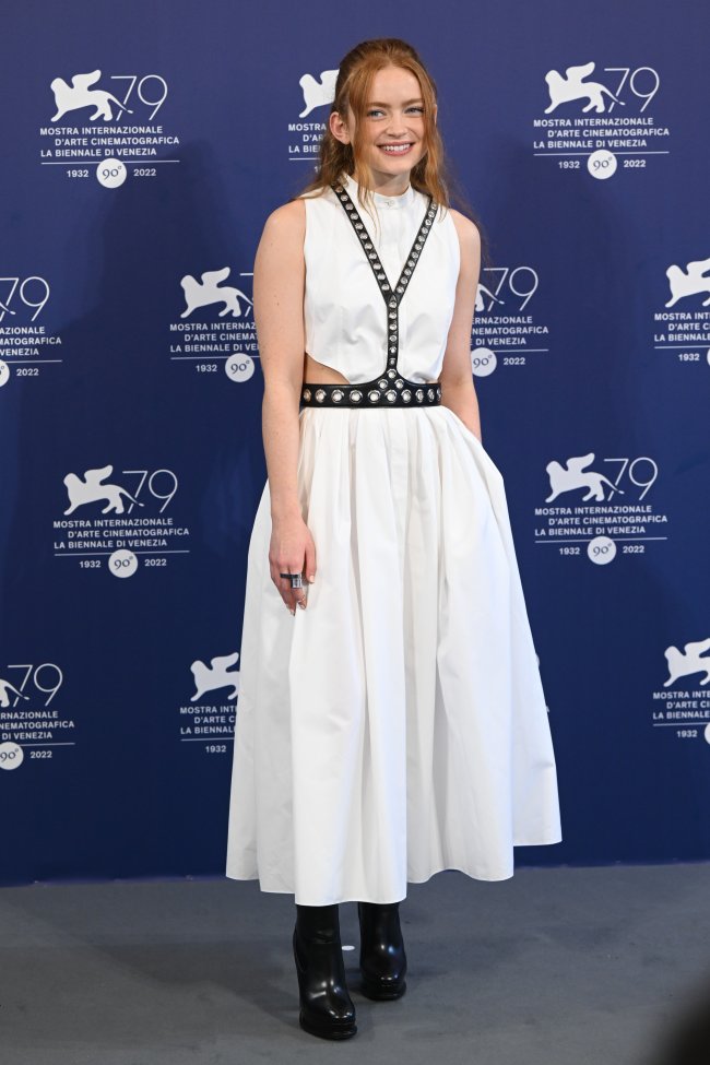 sadie-sink-no-festival-de-veneza Foto do look da atriz Sadie Sink no Festival de Veneza. Ela usa um vestido branco longo com detalhe de cinto preto e bota de salto alto preta. Ela olha para a câmera e sorri levemente.