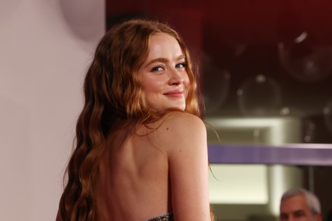 Foto da atriz Sadie Sink no tapete vermelho do Festival de Veneza. A foto é um close no rosto dela, ela está de perfil, olha para a câmera e sorri levemente.