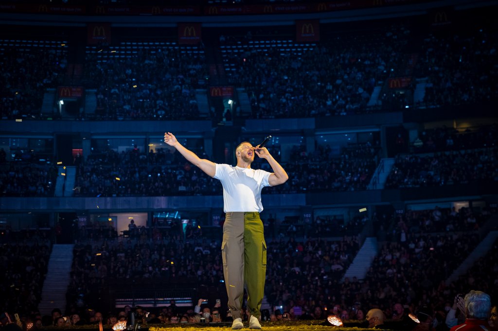 Dan Reynolds do Imagine Dragons se apresentando em show; ele está no palco com um dos braços levantado e segurando um microfone na outra mão enquanto canta