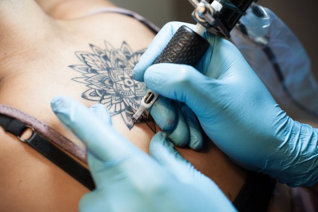 8 pontos para considerar antes de fazer sua primeira tatuagem Mão segurando um equipamento de tatuagem e fazendo uma mandala nas costas de uma jovem mulher