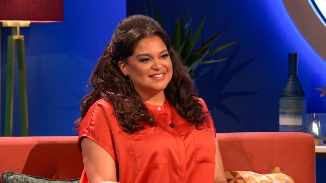 Michelle Buteau, apresentadora do The Circle US, sentada e sorrindo; ela usa uma blusa alaranjada e está sentada em um sofá com uma parede azul e uma planta ao fundo