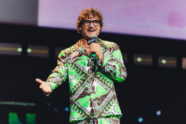 Pedro Pascal sorrindo enquanto segura microfone e fala no palco da CCXP22; ele usa um terno verde com branco