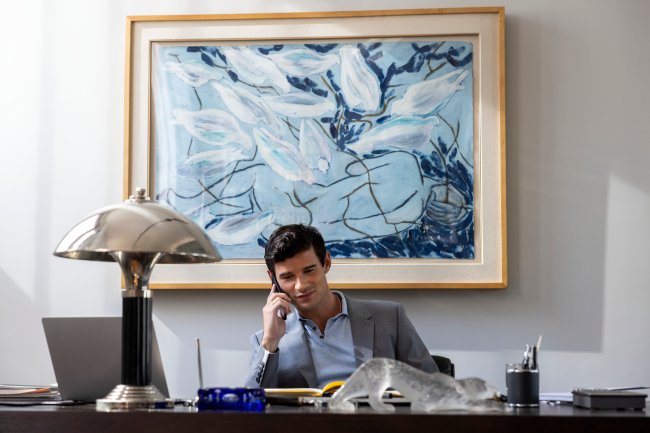 EIP_307_Unit_00529R Paul Forman como Nicolas De Leon em cena de Emily em Paris; ele está em um escritório com o celular no ouvido sorrindo levemente; ao fundo um quadro azul, branco e preto decora a parede