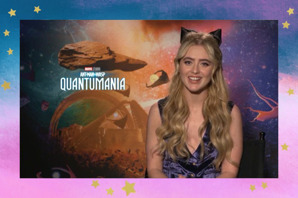 Kathryn Newton em entrevista de Homem-Formiga e a Vespa: Quantumania; ela está sorrindo em um fundo com elementos de rochas e pedras do mundo quântico com um céu estrelado em tons de laranja, vermelho, branco e roxo; a margem é um degradê de azul, roxo, branco, lilás e rosa; estrelas amarelas decoram a imagem