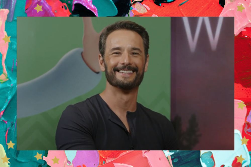 Rodrigo Santoro sorrindo enquanto olha para câmera; a margem é uma textura nas cores verde, roxo, vermelho, rosa, lilás e vermelho; estrelas amarelas decoram a imagem