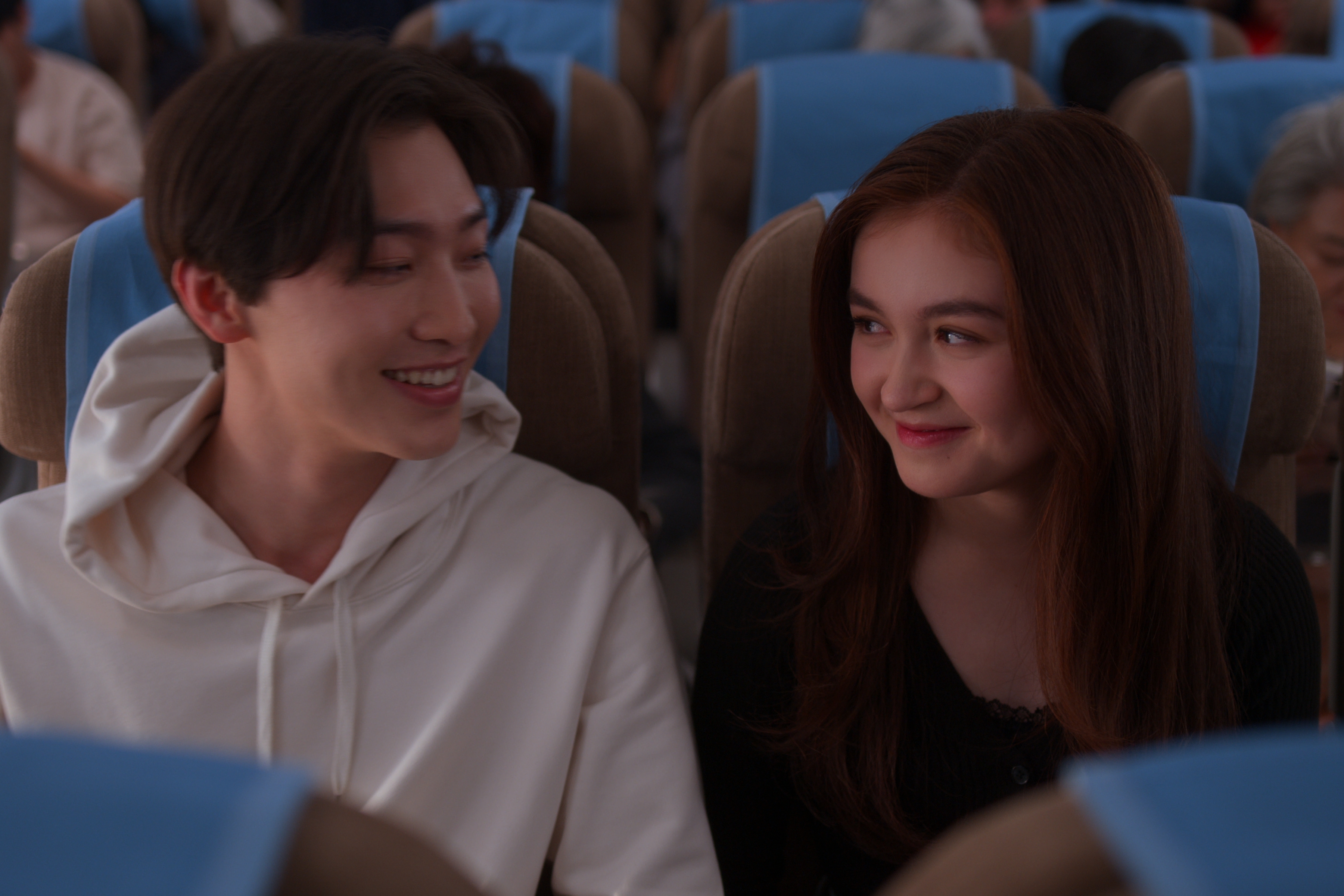 Sang Heon Lee como Min Ho e Anna Cathcart como Kitty Song Covey em Com Carinho, Kitty; eles estão em um avião sorrindo enquanto se olham