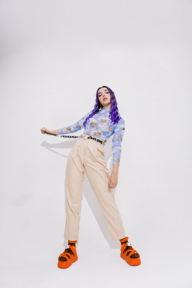 ana-xisde-sobre-moda Fundo branco, na frente Ana Xisdê aparece em pé com o corpo um pouco reclinado para a direita. Ela tem o cabelo roxo e veste uma blusa de manga comprida azul, um cinto escuro, uma calça bege e uma sandália laranja com meia.