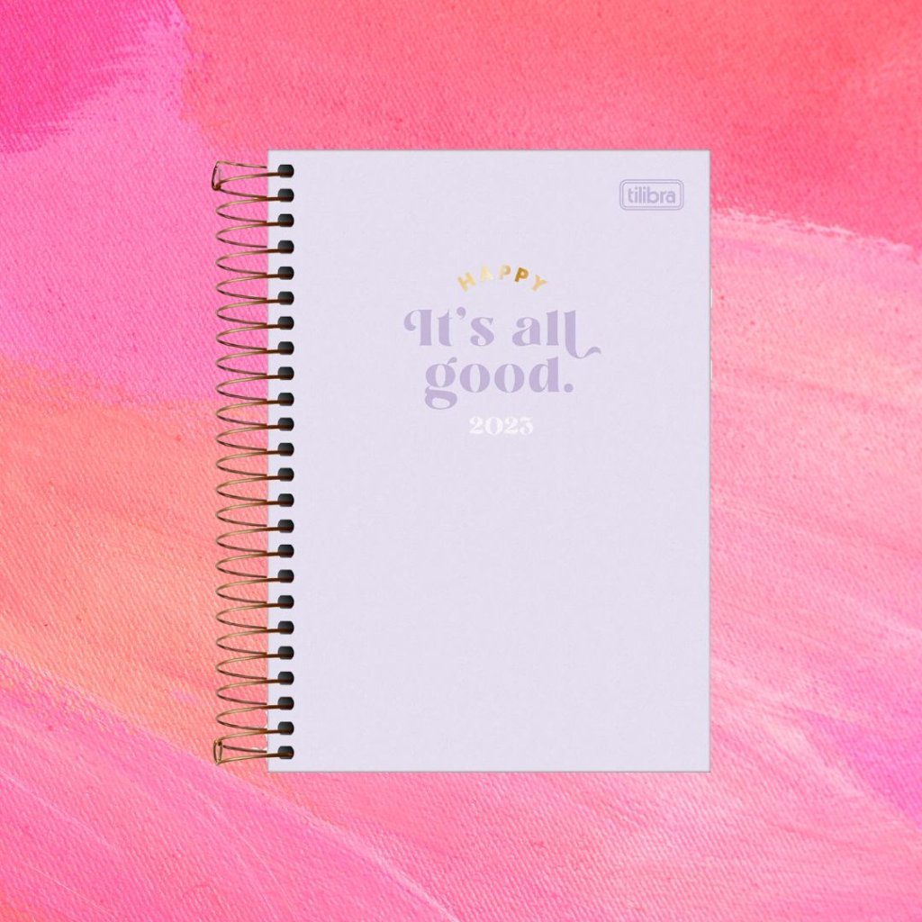 Planner lilás com texto "Happy/It's All Good/2023" em dourado, roxo e branco; o fundo é uma textura em tons de rosa e branco