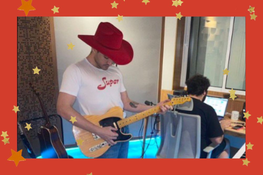 Foto do cantor Jão em estúdio. Ele está tocando guitarra e vestindo um chapéu vermelho e uma camiseta com escrito SUPER. Fundo vermelho com estrelas douradas