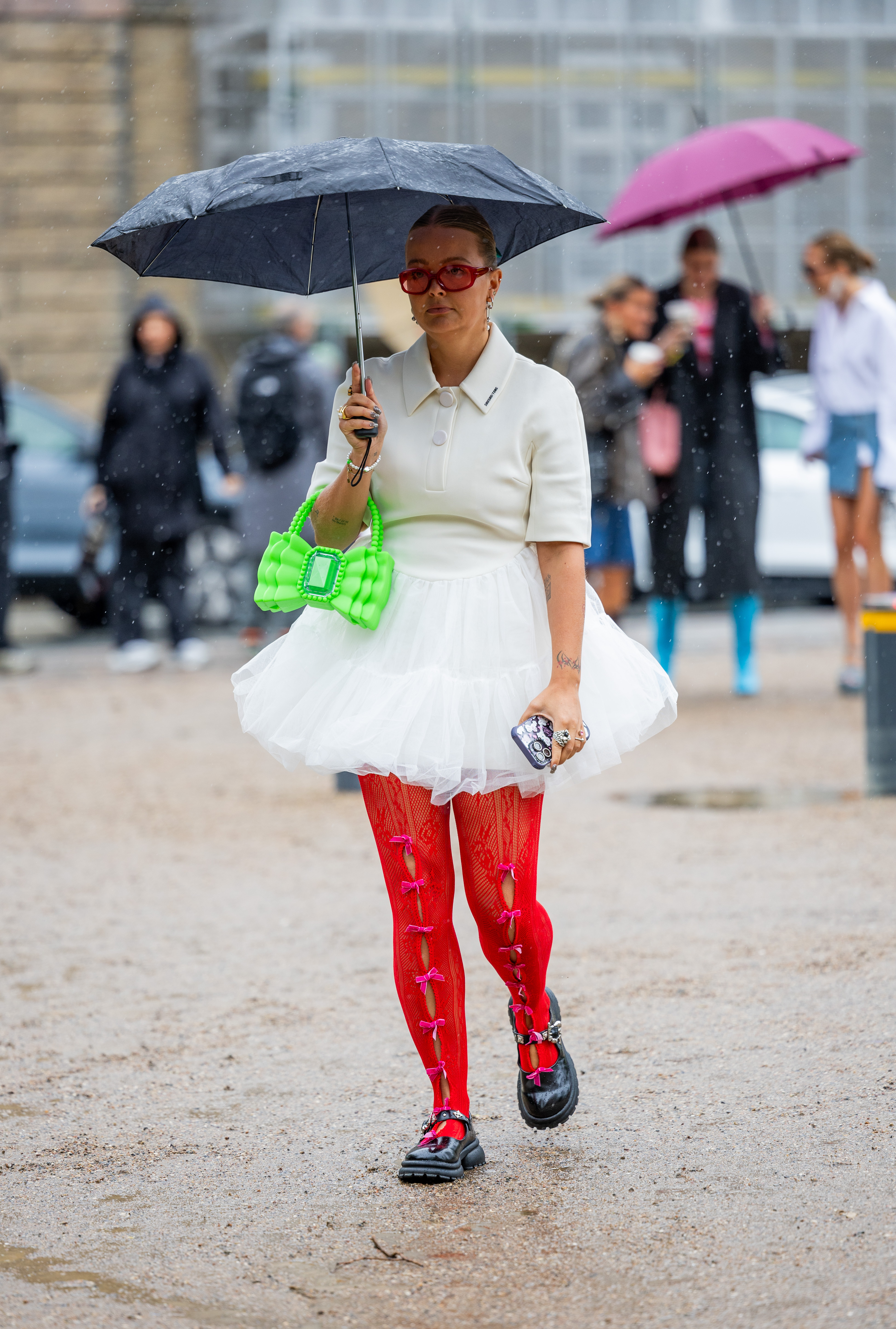 Street Style – Day 3 – Copenhagen Fashion Week Spring/Summer 2024 Look da semana de moda de Copenhagen na temporada de primavera-verão 2024 com blusa e saia off white, bolsa verde neon, meia-calça vermelha com laços e sapato preto