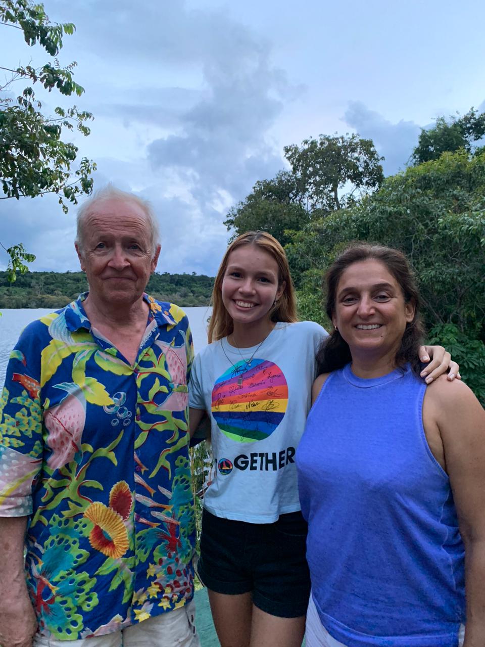 Doi lado esquerdo um senhor está vestindo uma camisa colorida, no meio uma garota loira de camiseta branca e do lado direito uma mulher de cabelos morenos e de camiseta azul