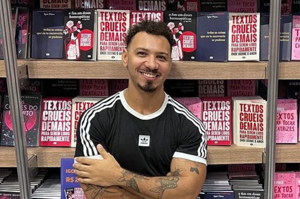 Igor Pires posando para foto sorrindo com os braços cruzados na frente do corpo; ele está de encostado em uma estante com prateleiras na cor marrom vários de seus livros expostos