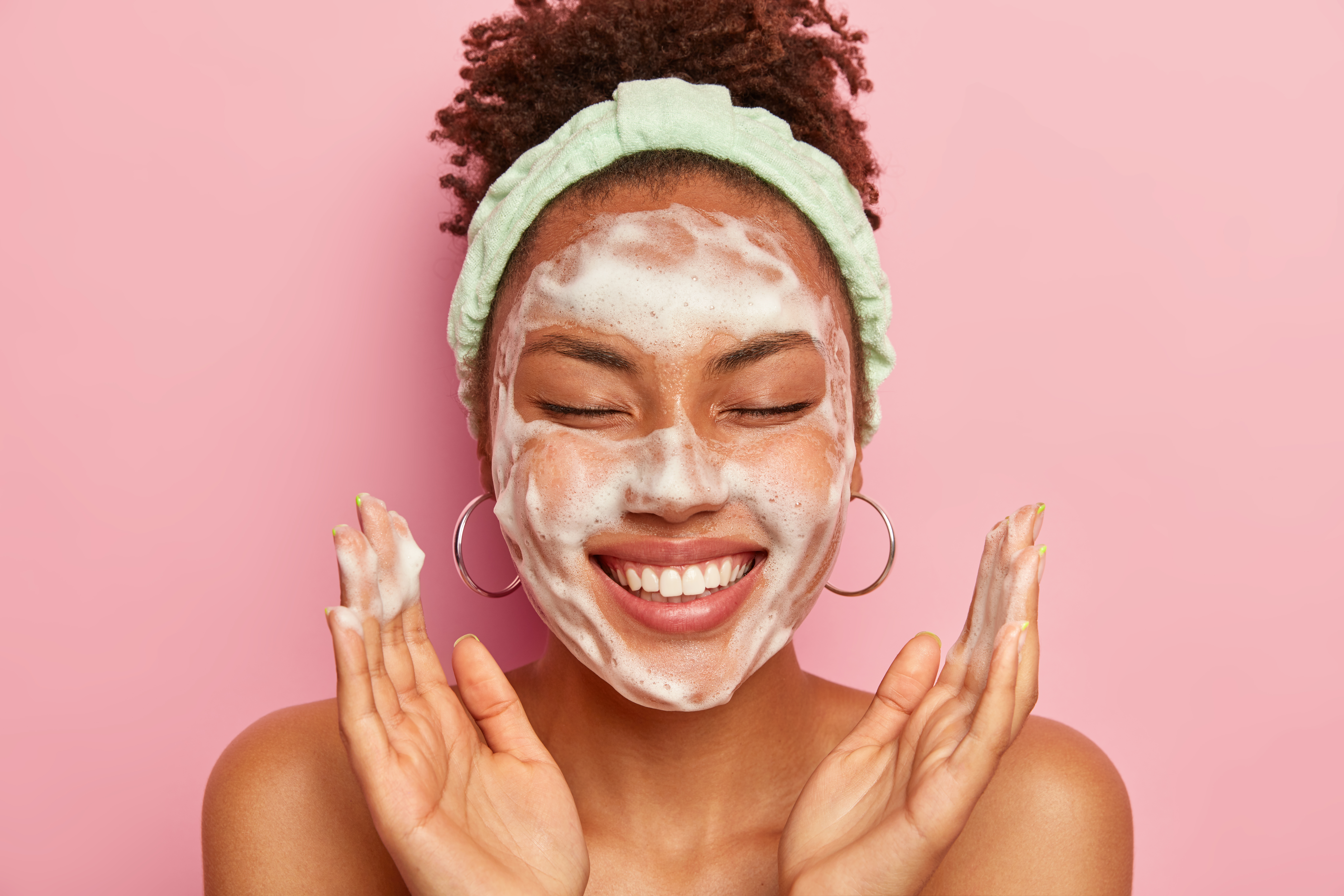 Image of happy ethnic woman raises palms over face, keeps eyes closed, shows white teeth, uses cleansing foam for skin care, gets real pleasure, wears headband and earrings, stands topless alone Garota com olhos fechados fazendo skincare com sabonete facial no rosto