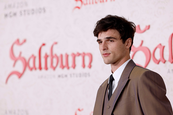 Jacob Elordi posando para foto com expressão séria; ao fundo está um backdrop com o nome do filme Saltburn em vermelho em um fundo branco