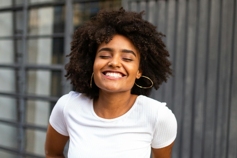 Menina de cabelo crespo sorrindo de olhos fechados usando blusa branca e brincos de argola