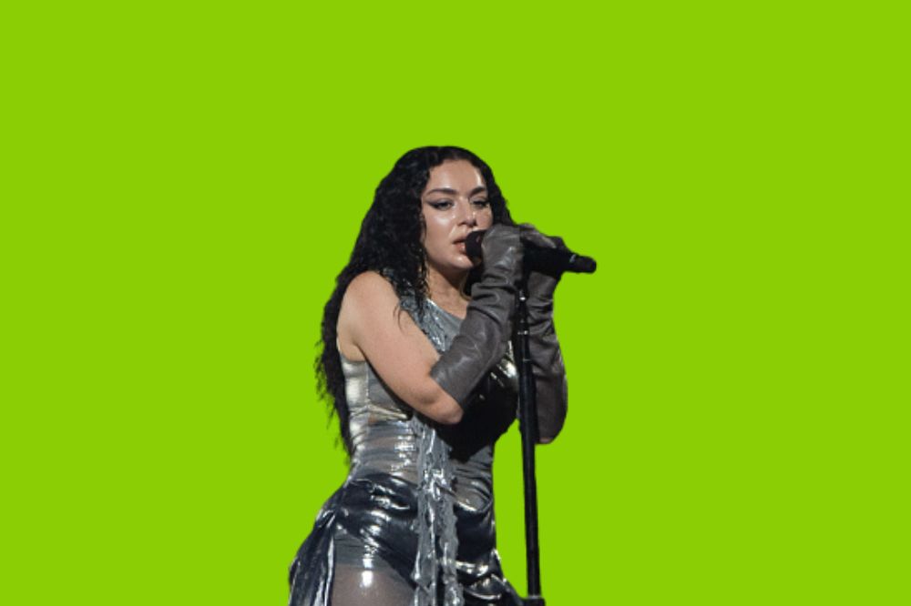 Foto da cantora Charli XCX cantando com roupa prata. Fundo verde no mesmo tom da capa do álbum "BRAT"