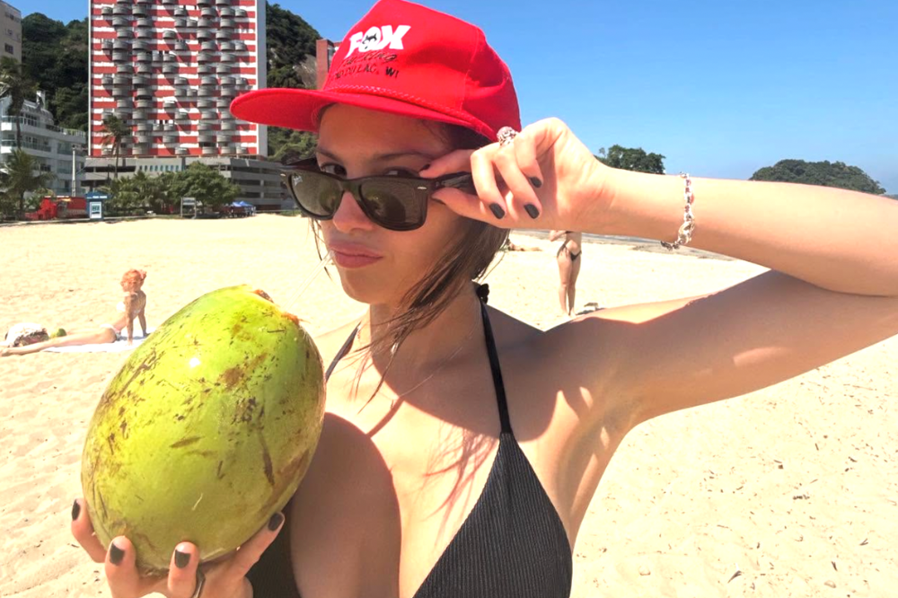 Olivia Rodrigo em praia brasileira com boné vermelho, óculos escuros e coco.