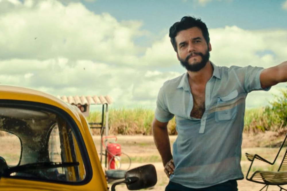 Wagner Moura aparece do lado de um carro em um espaço aberto