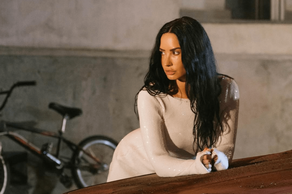 Demi Lovato usa um vestido branco cintilante e está apoiada sobre um carro olhando para a esquerda